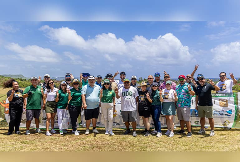 A large group of people associated with golf and UOG hold pose for a group photo outdoors, some holding up Triton hand sign
