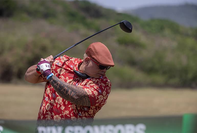 Man in red button up shirt and hat prepares to swing golf club outdoors