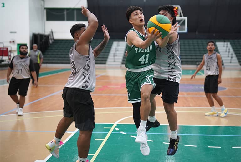 two players in white jerseys flank and try to block player in green jersey with basketball