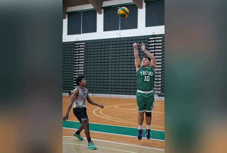 UOG Tritons player #10 makes jump shot from afar with opponent flanking.